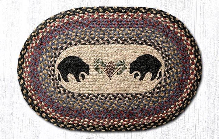 "A 20"x30" Oval Patch Braided Jute Rug featuring black, tan, cream, burgundy, and gray. Center of rug features two black bears standing facing one another with one paw up each. A sprig of pine with green needles and a brown pinecone is in the middle of the bears."